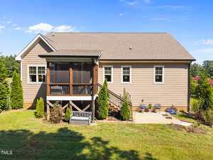 A single-story house with tan siding and a brown shingle roof. There is a screened porch with stairs, several windows, a small bench, and a seating area with two chairs and a table on a grassy lawn.