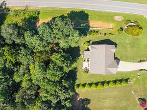 Aerial view of a single-story house with a gray roof, a curved driveway, and a large lawn. The property is bordered by dense trees on one side and a road at the top of the image.