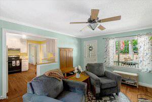 A cozy living room with light blue walls, a ceiling fan, two gray armchairs, a wooden coffee table with a flower vase, and a window with floral curtains. The kitchen and dining area are visible in the background.