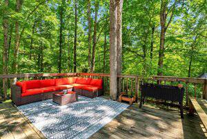 A wooden deck in a forest with a red cushioned sectional sofa, a small coffee table, a patterned outdoor rug, a black planter, and wooden railings surrounded by tall green trees.