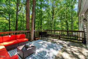 A wooden deck with a red outdoor sectional sofa, patterned area rug, small black grill, and trees surrounding the space. Sunlight filters through the leafy green canopy, creating a bright, inviting atmosphere.