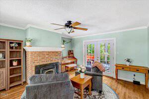 A living room features light green walls, a brick fireplace with a wood stove, two gray armchairs, a wooden side table, a piano, French doors, a wooden desk, and hardwood floors. Plants and decor accent the space.