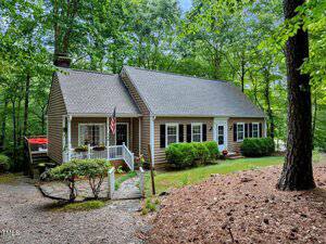 A brown, single-story house with white trim sits among tall trees at 160 Shadowbrook Drive in Henderson. The home has a front porch with an American flag, shrubbery lining the front, and a gravel path leading to the entrance.