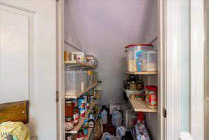 A small, narrow pantry with shelves on both sides holding various food items in jars, cans, and plastic containers. The floor stores bags and bottled water. The walls are painted white.