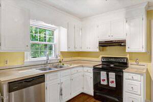 A kitchen with white cabinets, a black electric stove, a stainless steel dishwasher, a double sink under a window, and yellow walls. Two towels hang on the oven handle. Greenery is visible through the window.