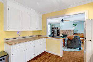 A kitchen with yellow walls, white cabinets, and wood flooring. A pass-through window opens to a living room with a brick fireplace, gray chairs, and bookshelves. A ceiling fan hangs in the living area.