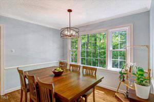 Dining area with a wooden table and six matching chairs, a bowl of fruit at the center, a modern light fixture above, large windows showing greenery outside, and a plant stand with various plants by the window.