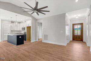 Open-concept modern kitchen and living area with light walls, wood-style flooring, a black kitchen island, stainless steel appliances, large ceiling fan, and a wooden front door with glass panel.