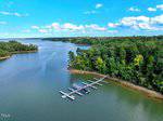 A dock with several boat slips extends from a wooded shoreline into a calm, wide lake. The water reflects the blue sky with scattered clouds, and dense green trees line the shore.
