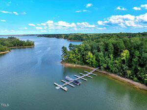 A dock with several boat slips extends from a wooded shoreline into a calm, wide lake. The water reflects the blue sky with scattered clouds, and dense green trees line the shore.