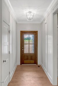A hallway with white paneled walls and wood flooring leads to a wooden front door with frosted and clear glass panels. A modern ceiling light fixture is above the entryway.