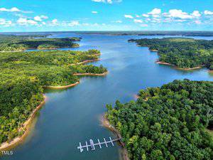 Aerial view of a large lake surrounded by dense green forests, with a dock extending into the water. Small islands and peninsulas are visible under a partly cloudy blue sky.