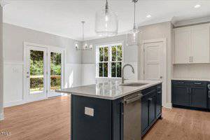 Modern kitchen with light wood floors, white walls, and navy blue lower cabinets. Features a white island with a sink, stainless steel fixtures, and large windows overlooking a green outdoor area.