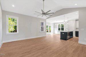 Spacious, modern open-concept living area with light wood floors, large ceiling fan, neutral walls, and a kitchen featuring a central island, pendant lights, and stainless steel appliances. Large windows provide natural light.