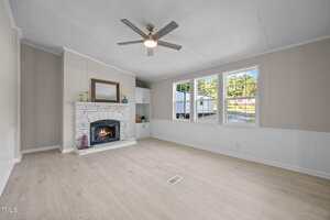 A spacious, empty living room with light wood flooring, a white brick fireplace, a ceiling fan, and three large windows letting in natural light. There are built-in shelves next to the fireplace.