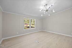An empty room with light wood flooring, beige walls, a modern ceiling light fixture, and a double window offering a view of a grassy yard with trees and shrubs outside.