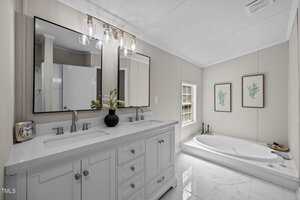 Modern bathroom with a double-sink vanity, two mirrors, and a countertop clock and vase. There is a bathtub by a window, two botanical art prints on the wall, and white tile flooring.