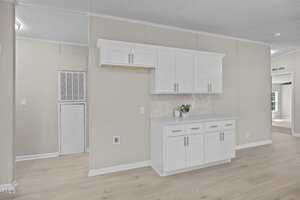 A section of a modern interior with light wood floors, beige walls, white upper and lower cabinets with silver handles, a small countertop, a potted plant, and a vented utility area on the left.