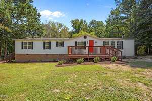 A single-story manufactured home at 261 Ragland Lane, Henderson, features white siding, a brick foundation, red front door, and wooden porch, surrounded by grass, small shrubs, and trees under a clear blue sky.