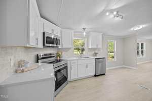 Modern kitchen with white cabinets, stainless steel appliances, a large window above the sink, light wood flooring, and neutral walls. The kitchen opens into a bright living space with large windows.