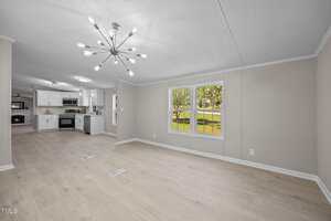 A modern, unfurnished living and kitchen area with light wood floors, beige walls, a large window overlooking a grassy yard, and a contemporary ceiling light fixture. The kitchen has white cabinets and stainless steel appliances.