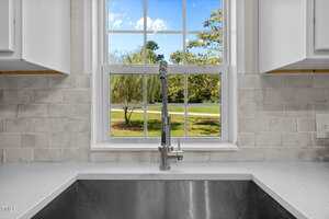 A stainless steel kitchen sink with a modern faucet is set against a white tile backsplash beneath a window, which shows a clear blue sky and green trees outside.