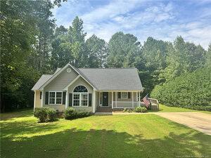 A single-story beige house with white trim and a front porch sits on a well-maintained lawn at 4138 GENEVA Drive in Franklinton. The gabled roof, American flag by the porch, and surrounding trees complete this charming scene under a partly cloudy sky.