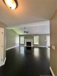 Spacious living room with dark hardwood floors, gray walls, white ceiling, and a ceiling fan with light. There is a white fireplace centered between two windows, and a glass door to the right.