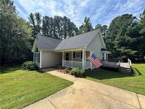 A single-story beige house with a gable roof, front porch, and ramp. An American flag is mounted on the porch. The house is surrounded by a grassy lawn and trees under a partly cloudy sky.