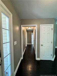 A hallway with dark wood floors, light gray walls, and white trim, featuring several white doors, a ceiling light fixture, and a glass panel door on the left side.