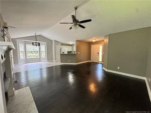 Open-concept living area with dark wood floors, adjoining kitchen with white cabinets, a dining space with tile flooring, ceiling fan, and a view of the front door and windows in the background.