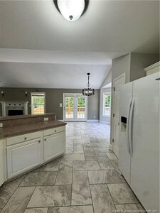 Spacious kitchen with white cabinets and a white refrigerator, opening to a living area with a fireplace and dining space. Large windows and double French doors provide natural light and access to a wooden deck.