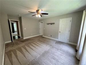 A clean, empty bedroom with light gray walls, beige carpet, a ceiling fan with lights, two white doors, a small wall shelf, and large windows with sheer curtains letting in natural light.