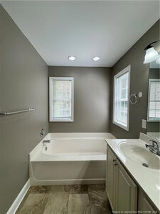 A bathroom with a white bathtub next to two windows, a towel bar on the wall, a sink with a counter and cabinet, a mounted light fixture, and beige tile flooring. The walls are painted gray.