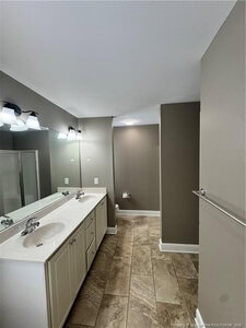 A bathroom with beige tiled floor, double white sinks with cabinets, a large mirror with two light fixtures above, and taupe-colored walls. A shower stall is partially visible on the left.