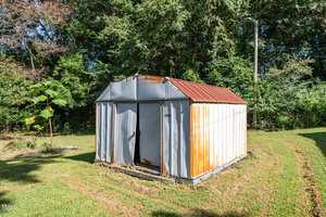 A metal storage shed with a rusted roof and sides stands on a grassy lawn, surrounded by trees and greenery in the background. The doors of the shed are closed.