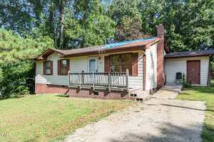 A single-story beige house with brown trim and a small front porch sits at 110 Lynnbank Estates Road, Kittrell. Part of the roof is covered with a blue tarp. The grassy yard features trees, greenery, and a concrete driveway.