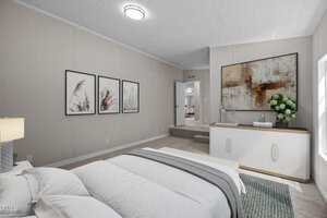 A bedroom with a neatly made bed, light-colored walls, framed art above the bed, a white dresser with a large abstract painting, and a window letting in natural light. The carpet is beige, and a lamp sits on the nightstand.