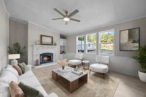 A living room with a white sofa, two white armchairs, a coffee table, a fireplace with a white brick surround, houseplants, large windows, neutral walls, and a ceiling fan with a light.