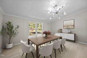 A dining room with light walls and floors, a wooden table with six white chairs, a modern chandelier, a sideboard with decor and artwork, a potted plant in the corner, and large windows letting in natural light.
