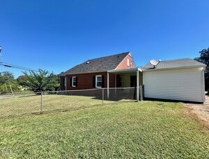 A one-story house with red brick and white siding, a dark shingled roof, small covered porch, and a fenced-in grassy yard under a clear blue sky. A satellite dish is attached to the side extension.