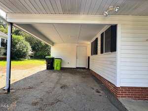 A covered carport with a white ceiling and siding, black shutters, brick trim, and two garbage bins near a white door. The ground is paved with some cracks and patches. Trees and grass are visible in the background.