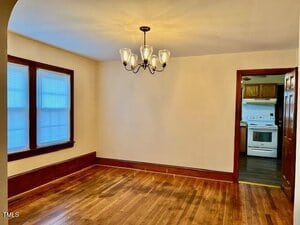 A dining room with hardwood floors, cream-colored walls, a dark wood trim, a window with blinds, and a six-light chandelier. An open doorway leads to a kitchen with wood cabinets and white appliances.