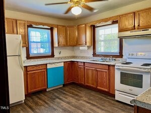 A kitchen with wood cabinets, white appliances including a stove and refrigerator, a blue dishwasher, double sink under two windows, light granite countertops, and dark wood flooring. A ceiling fan is on the white ceiling.