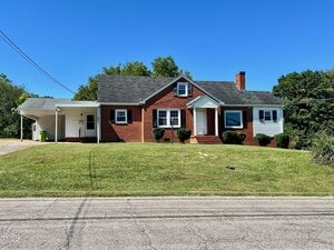A red brick and white siding single-story house with a black roof, a carport on the left, trimmed bushes, and a well-maintained lawn, set against a clear blue sky.