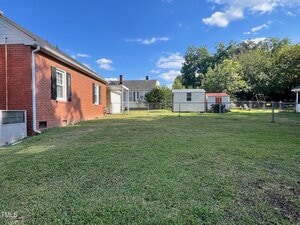 A fenced backyard with a well-maintained lawn, a red brick house on the left, two small sheds in the distance, trees along the back fence, and a blue sky with scattered clouds overhead.