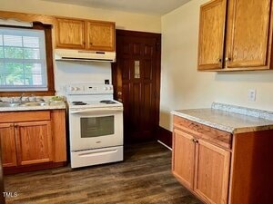A kitchen with wooden cabinets, a white electric stove with oven, a range hood, a double sink under a window, and a brown door. The countertops are light-colored, and the floor is dark wood.
