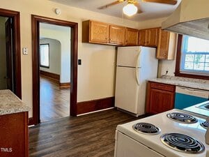 A kitchen with wood cabinets, white appliances including a refrigerator and stove, granite countertops, a window with blinds, and brown wood trim, opening into a room with hardwood floors.