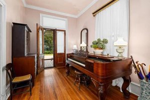 A small room with hardwood floors features a piano with a bench, a cabinet, a chair, a mirror, lamps, potted plants, and umbrellas. Double doors open to an outdoor area, letting in natural light through sheer curtains.