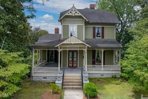 A two-story Victorian-style house with a covered front porch, decorative trim, gabled roof, and large windows. The house is surrounded by green shrubs and trees, with steps leading up to the front entrance.